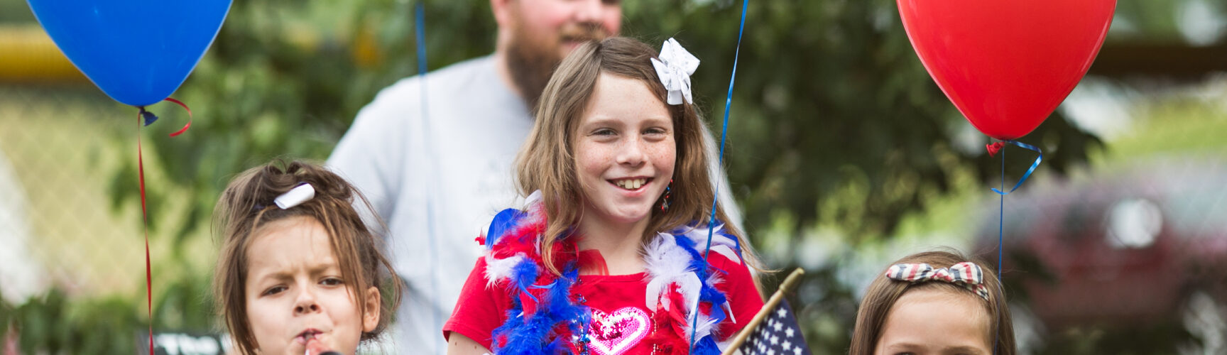Children in red white and blue patriotic outfits holding flags and balloons.